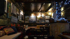 Un ambiente acogedor y con poca luz, con vigas de madera rústicas, un muro de piedra decorado con cuadros, y una mesa de madera con diversos objetos.