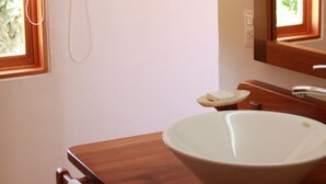 Un baño con tocador de madera, lavamanos blanco, espejo y ventana con persiana.