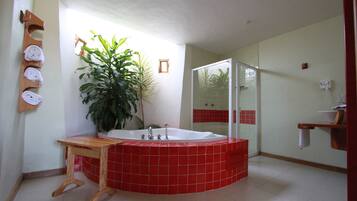 A bathroom with a red-tiled jacuzzi, a wooden vanity, a large potted plant, a glass-enclosed shower, and a towel rack with folded towels.