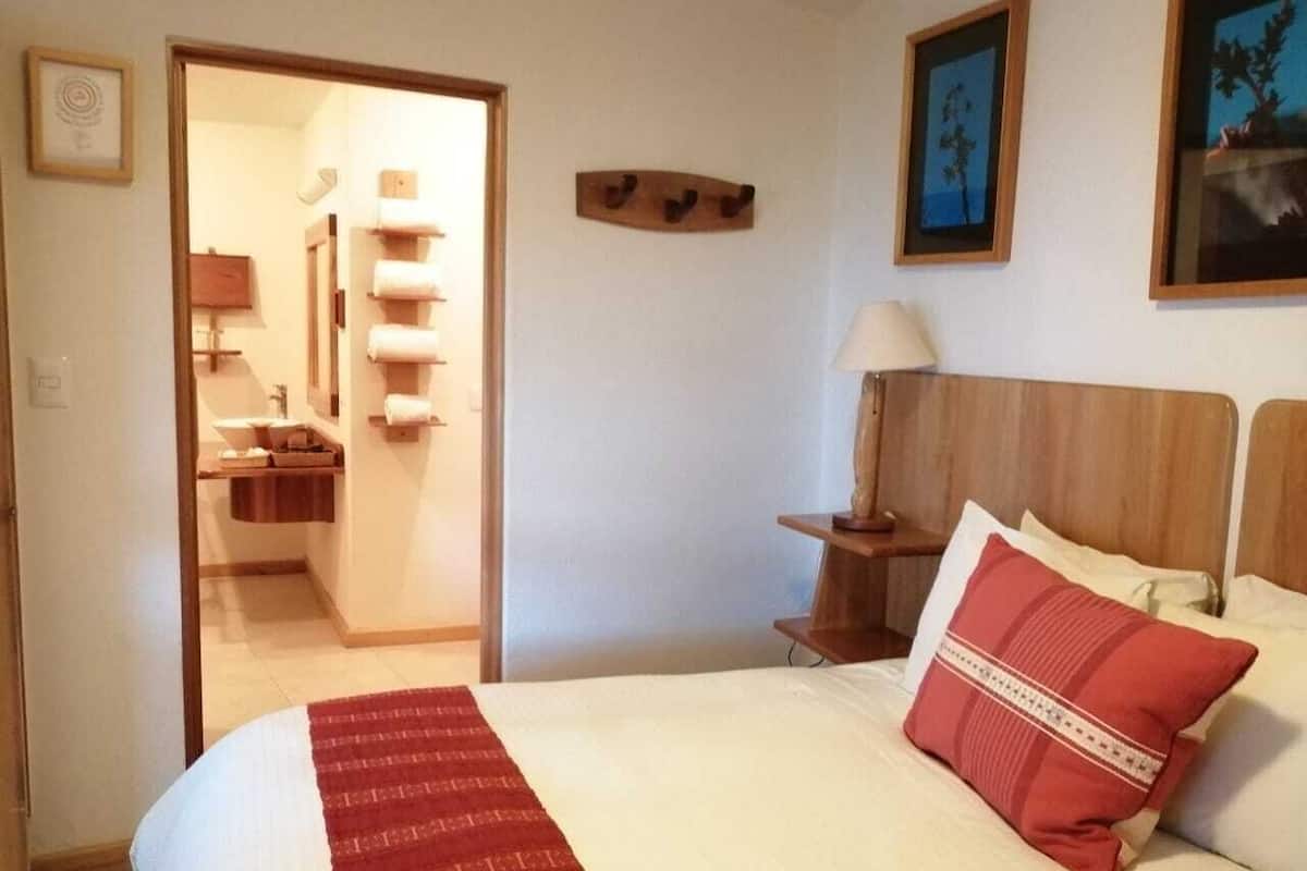 Hotellihuone, jossa on sänky, yöpöytä, lamppu, puinen pääty ja ovi, joka johtaa kylpyhuoneeseen.