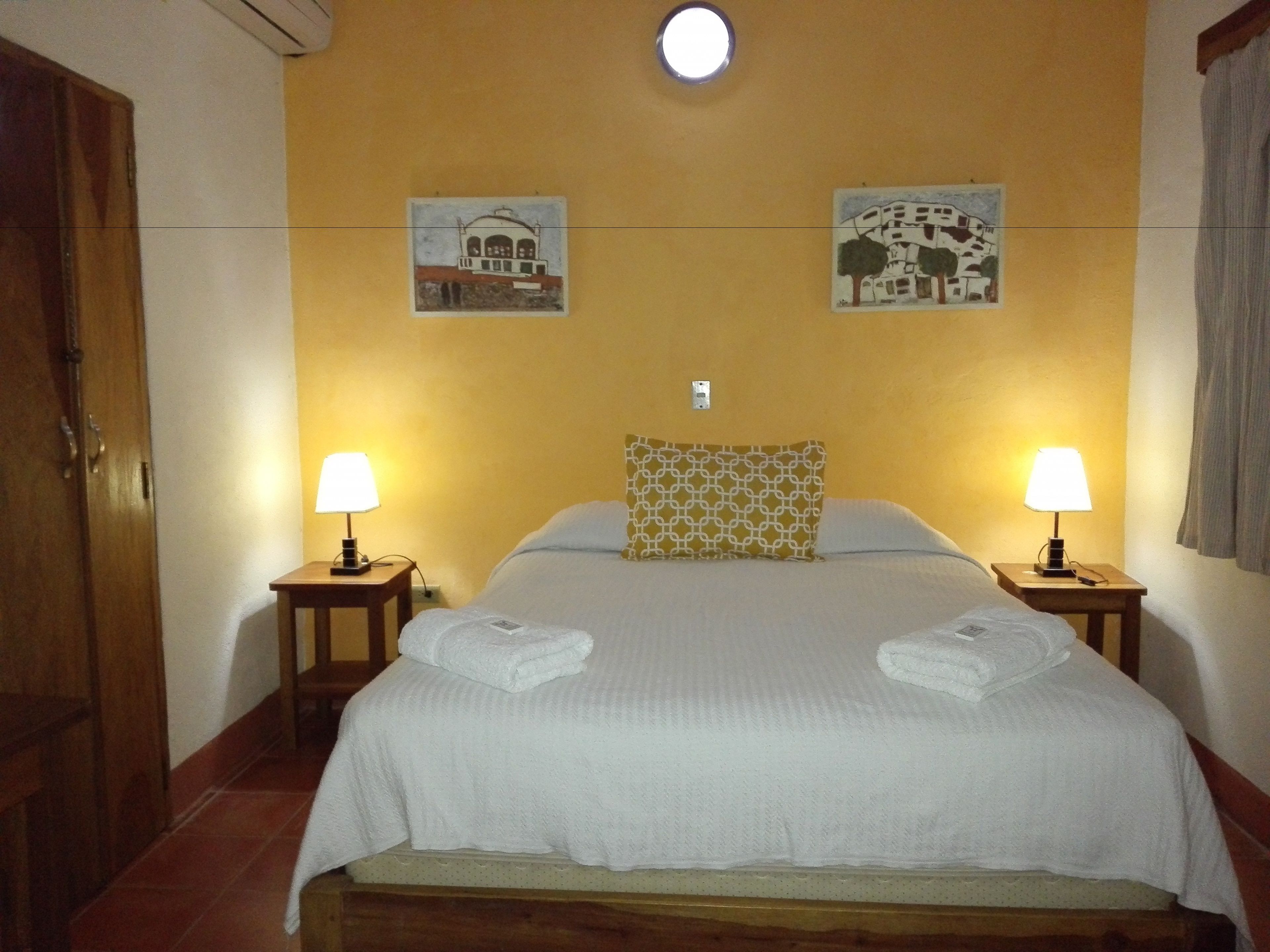 A hotel room with a bed, two bedside lamps, a wall-mounted light, and two wall art pieces.
