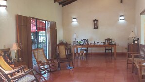 A spacious living room with a wooden floor, a dining table, and several wooden chairs. There is a large window with a view of greenery outside. - Hotel Casa Barcelona (Granada)