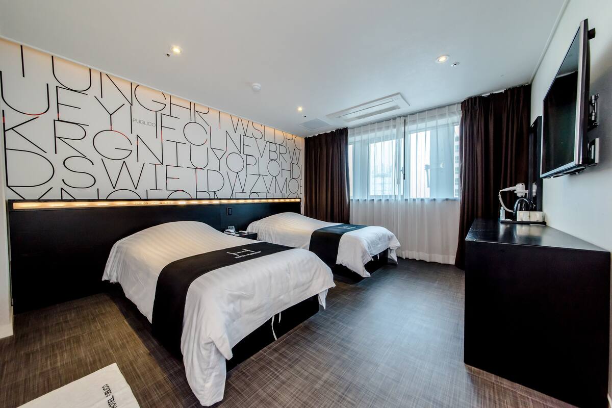 Habitación de hotel con dos camas, una pared grande con texto abstracto, un televisor de pantalla plana y un escritorio.