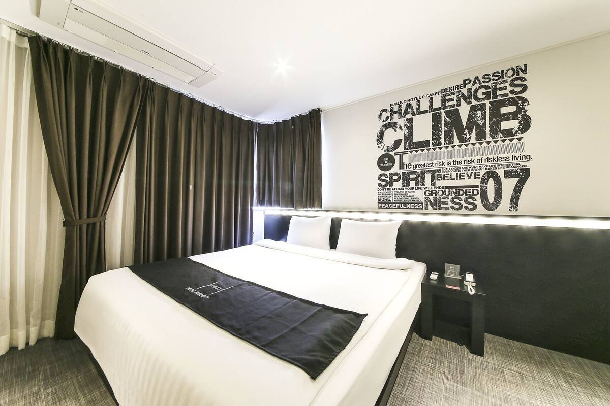 Habitación de hotel con una cama grande, un cabecero negro, una pared con texto motivacional y una ventana con cortinas.