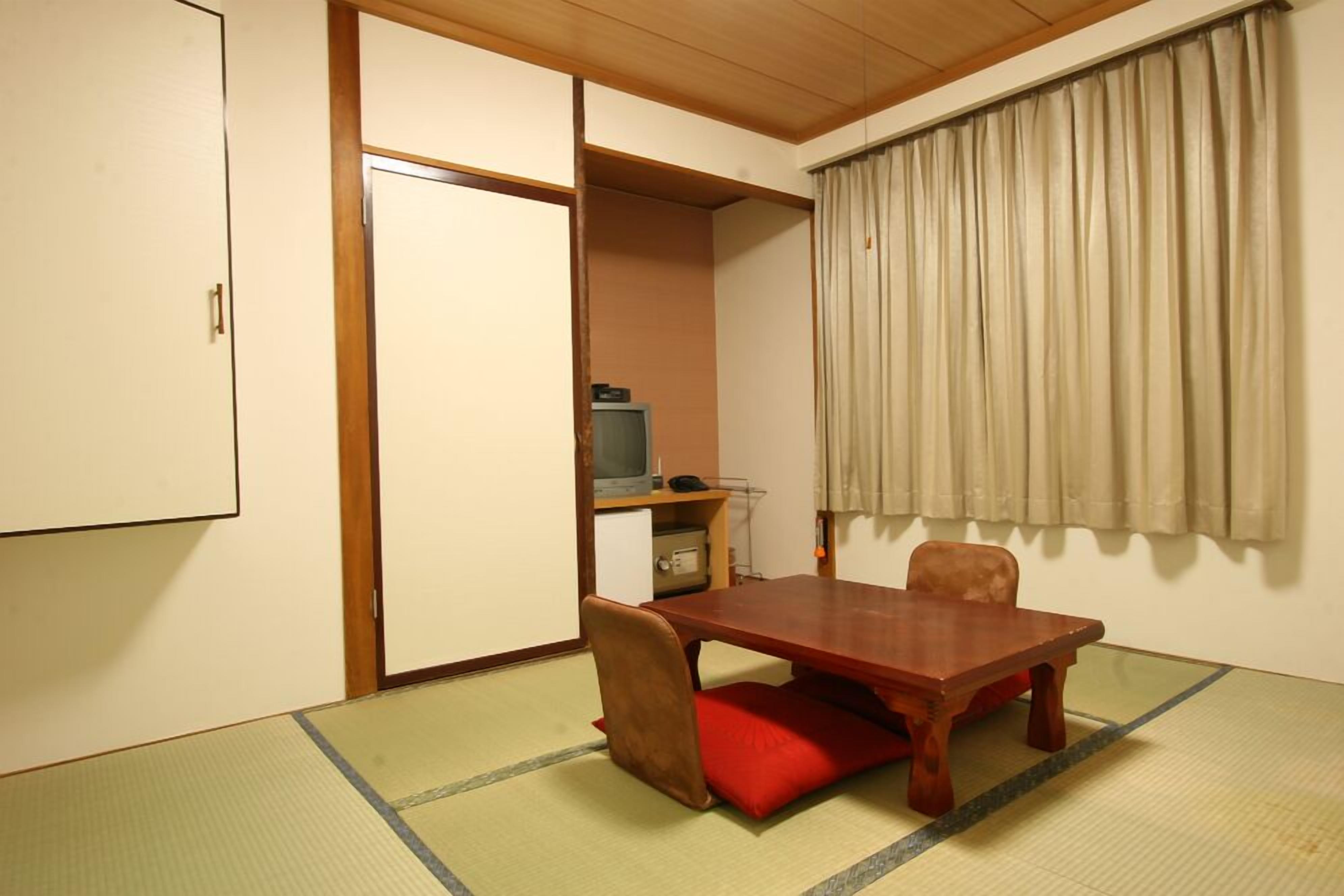 A room with tatami flooring, a wooden table, a red cushion, a television, and a sliding door.