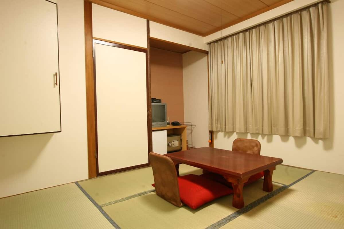 Ein Zimmer mit Tatami-Boden, einem Holztisch, einem roten Kissen, einem Fernseher und einer Schiebetür.