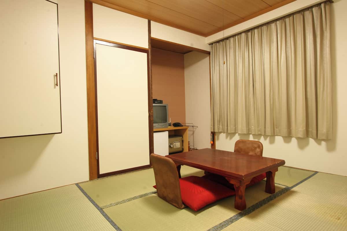 Una habitación japonesa tradicional con suelo de tatami, una mesa de madera, un cojín rojo, un televisor y una puerta corredera.