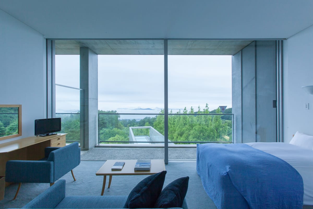 A modern hotel room with a large bed, a desk with a computer, a chair, and a view of the outdoors.
