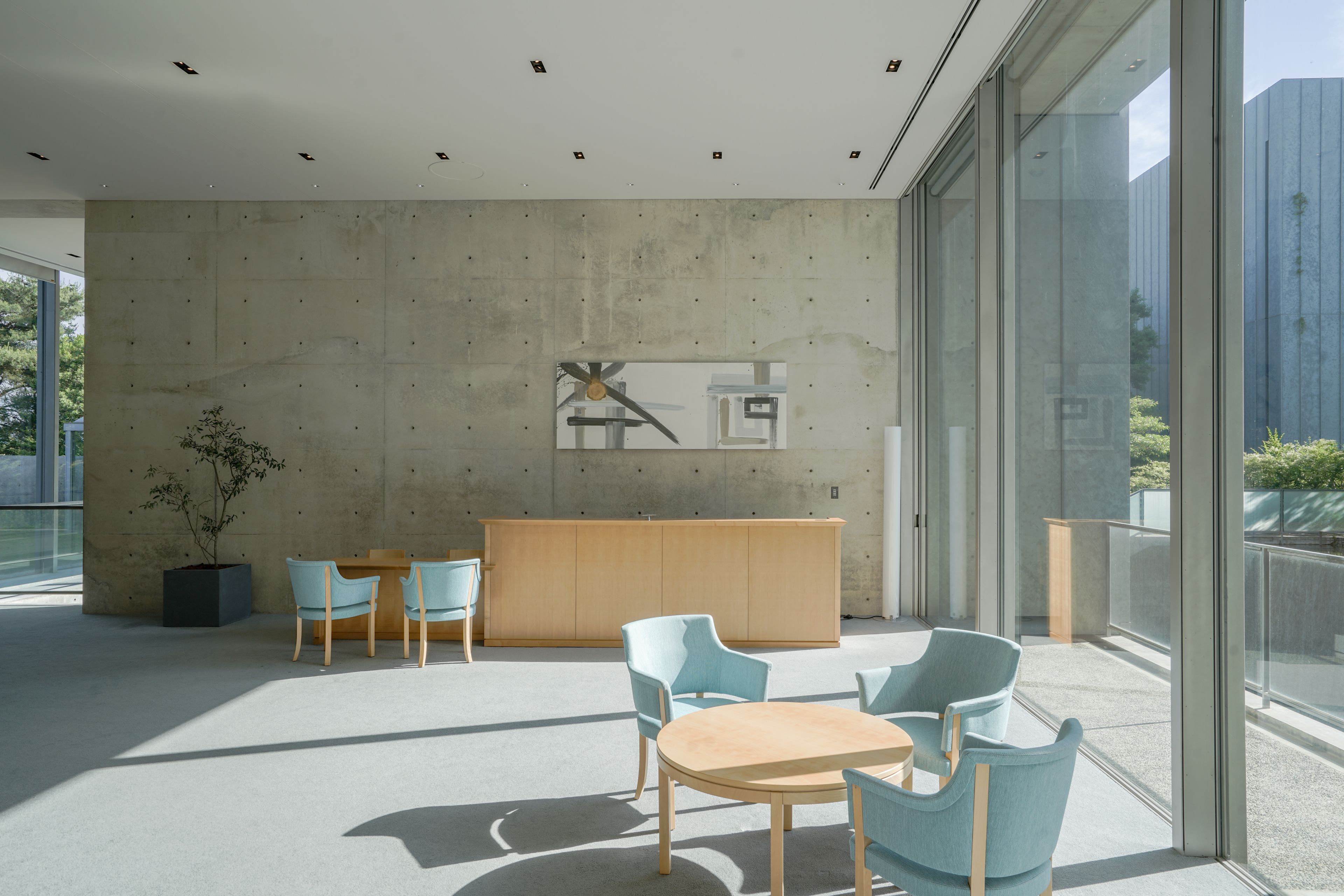 Um interior moderno com parede de concreto, balcão de recepção e cadeiras azul-claras ao redor de uma mesa de madeira.