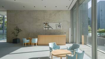 Um interior moderno com parede de concreto, balcão de recepção e cadeiras azul-claras ao redor de uma mesa de madeira.
