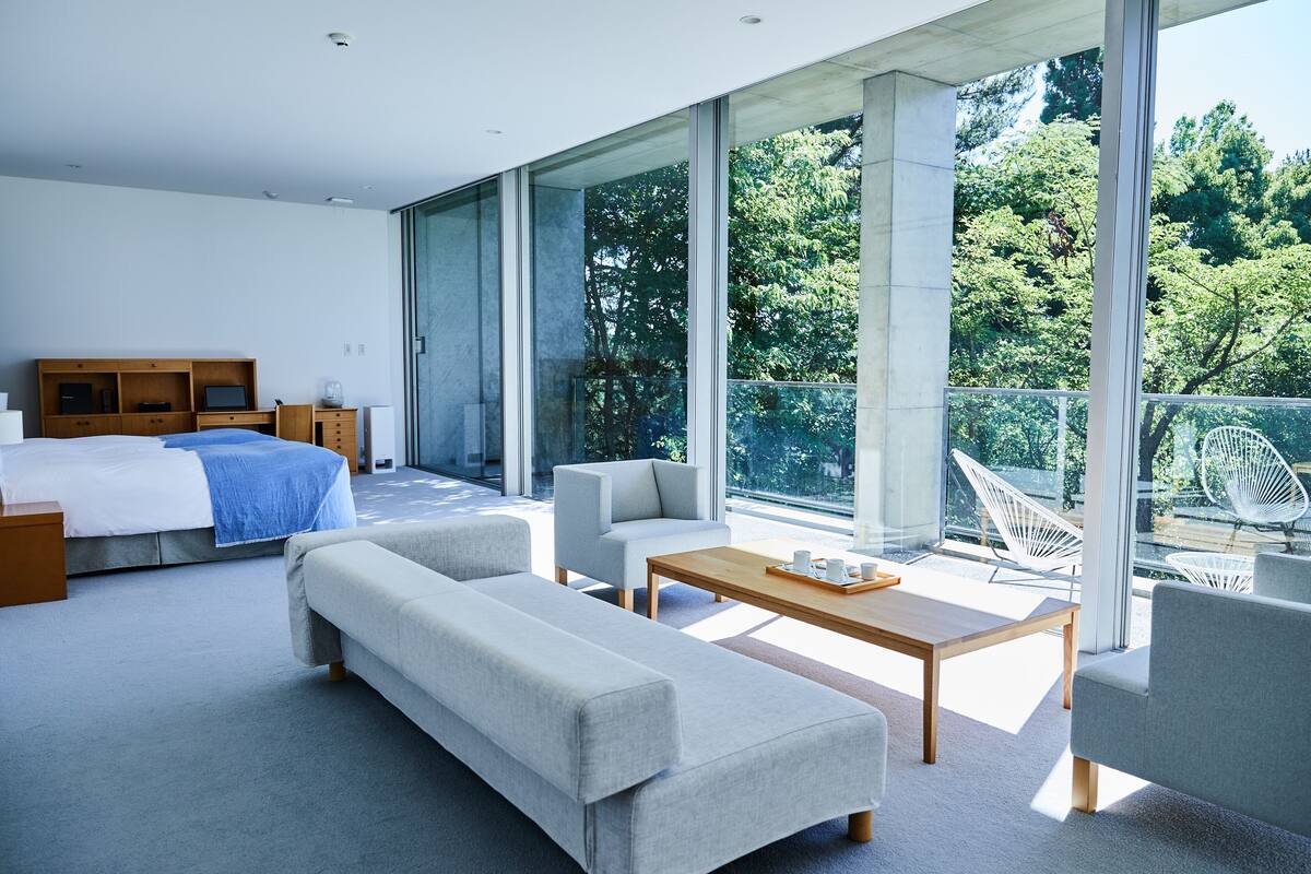 A modern bedroom with a large bed, a sofa, a coffee table, and a balcony with a view of trees.