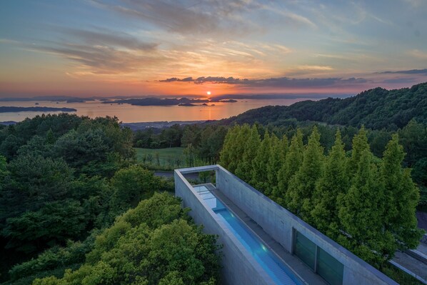 A modern building with a swimming pool, surrounded by lush greenery and overlooking a body of water during sunset. - SETOUCHI RETREAT AONAGI by Onko Chishin (Matsuyama)
