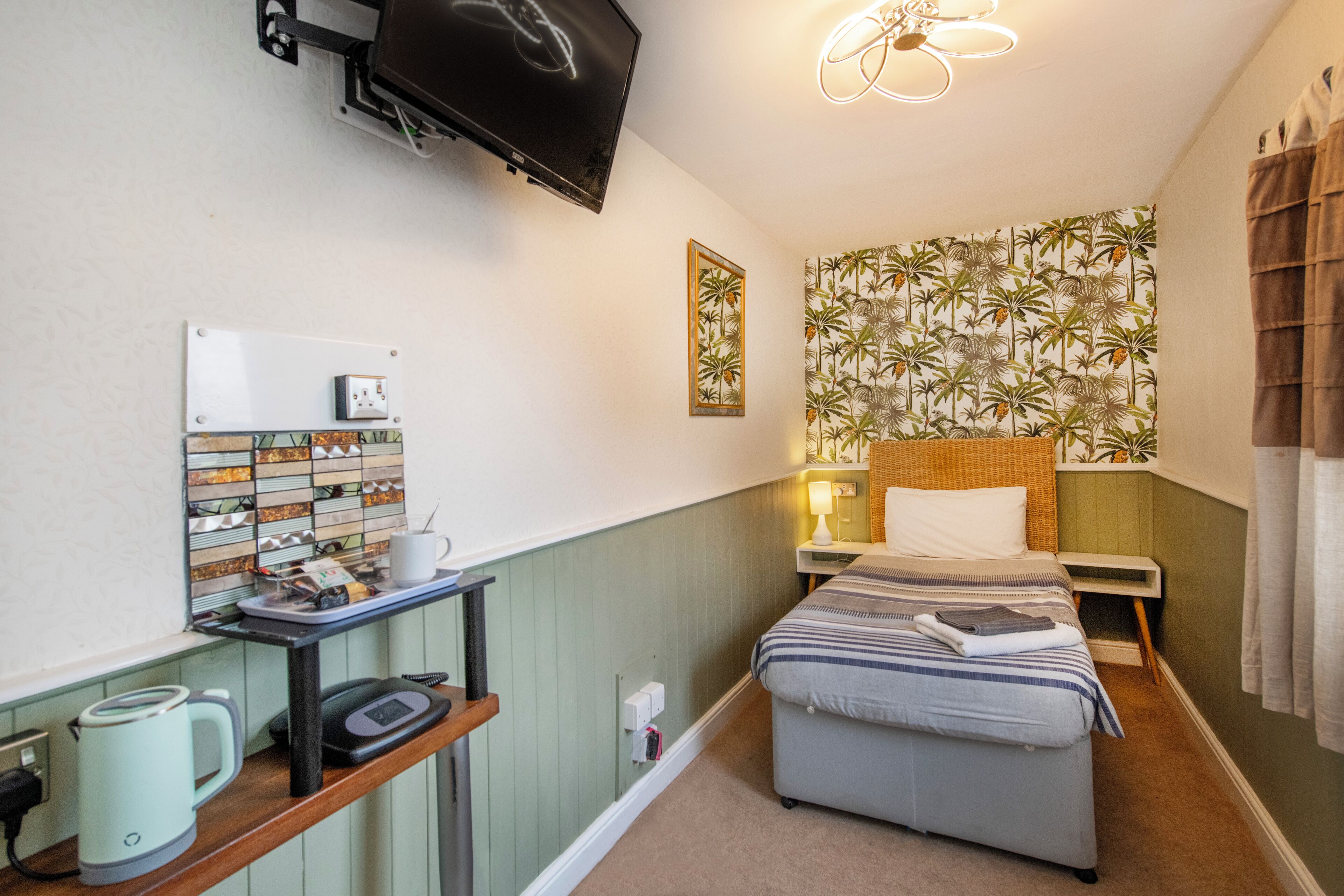 A compact hotel room with a bed, a desk with a kettle, a television, and a patterned wallpaper.