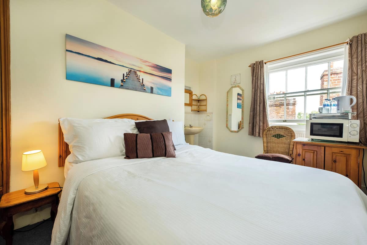 A hotel room with a large bed, a bedside lamp, a painting of a sunset, a window with curtains, a microwave, and a sink.