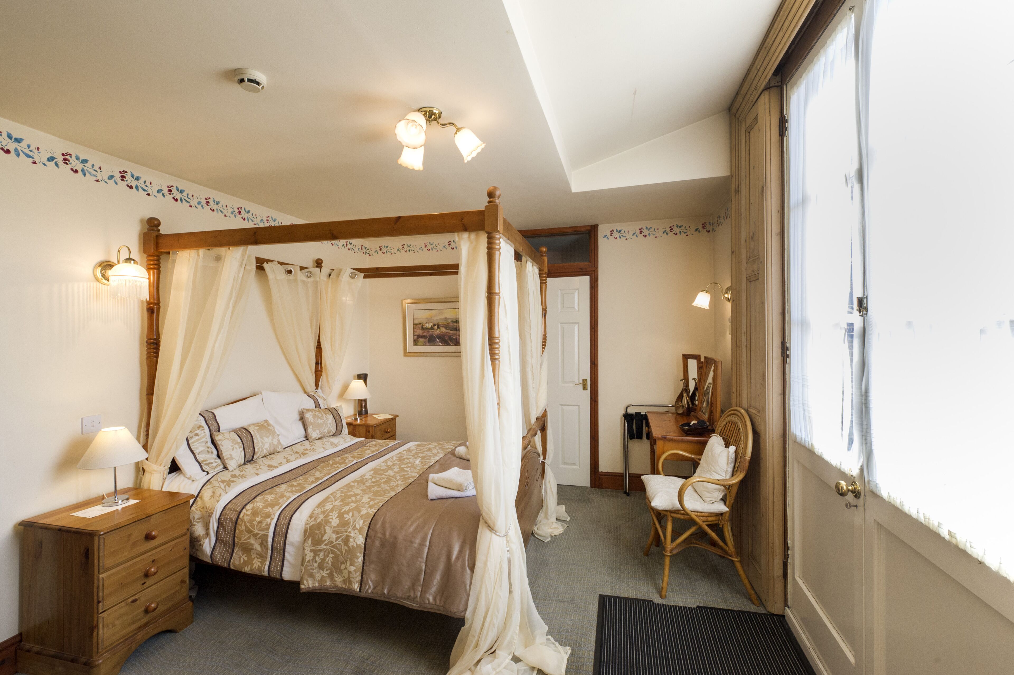 A four-poster bed with a canopy, a bedside table with a lamp, a chair, and a door leading to another room.