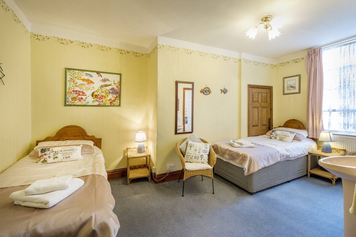 A hotel room with two beds, a chair, a bedside table with a lamp, a window with curtains, and a wall with a floral border.