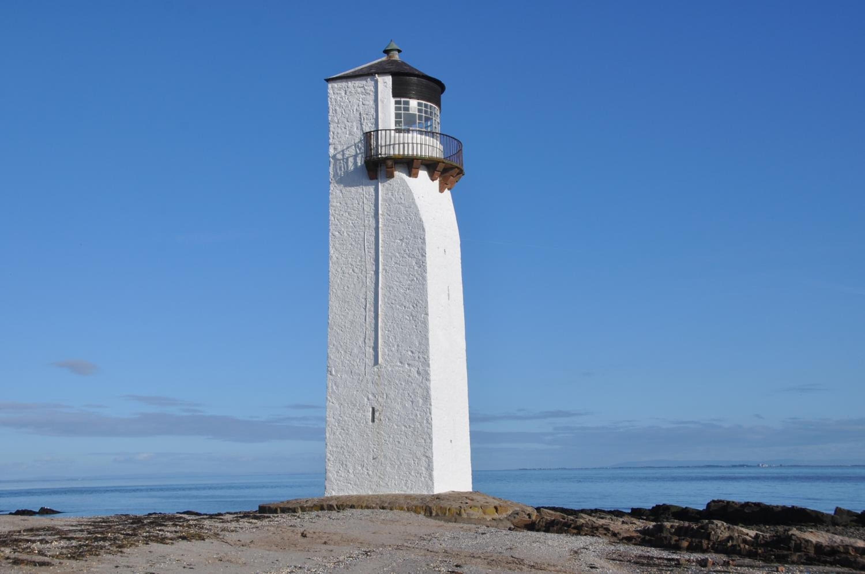 A tall, narrow lighthouse with a white exterior and a dark top, situated on a rocky shoreline with the sea in the background.