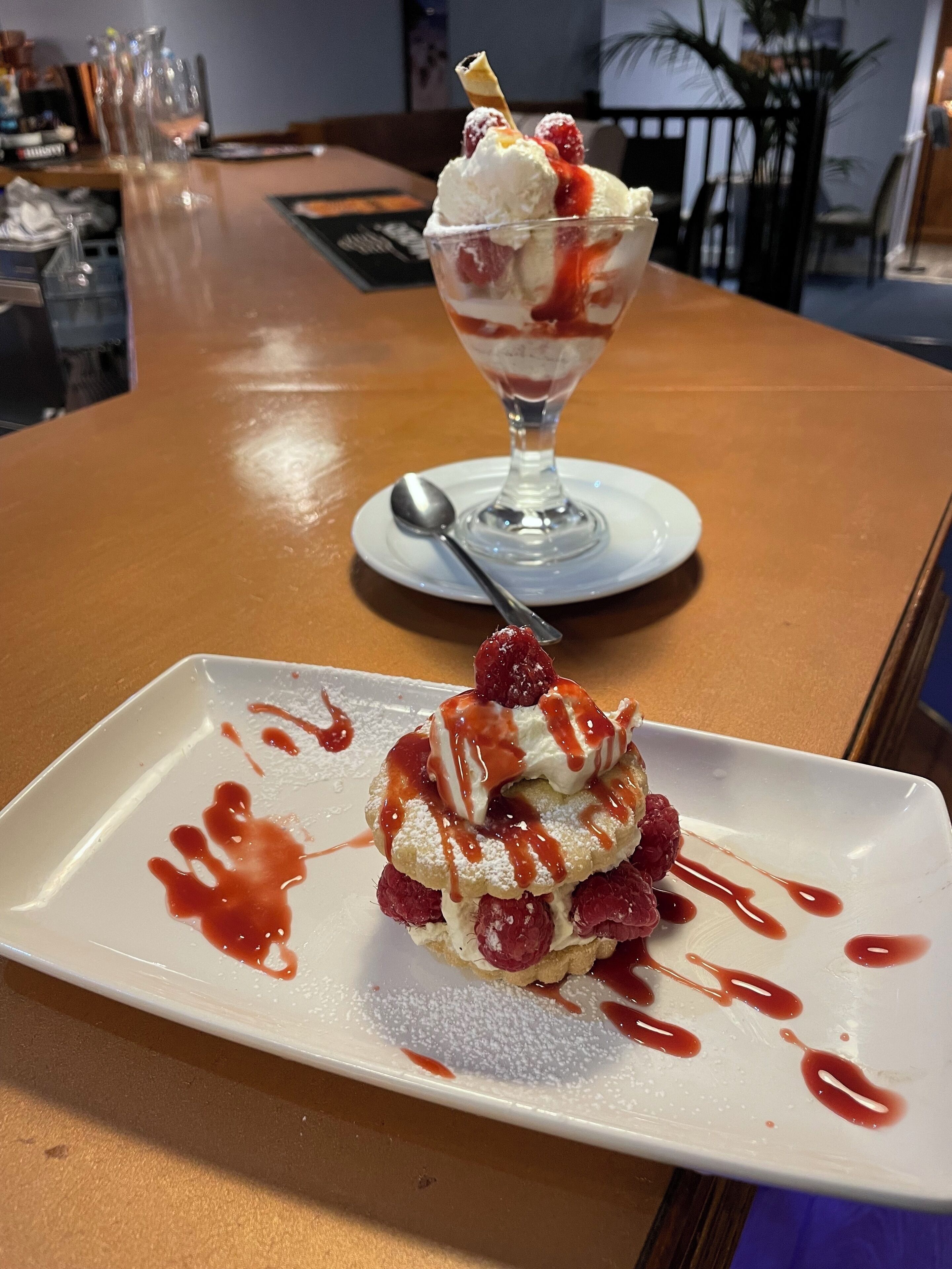A dessert on a white plate with a spoon, and a dessert glass in the background on a wooden table.
