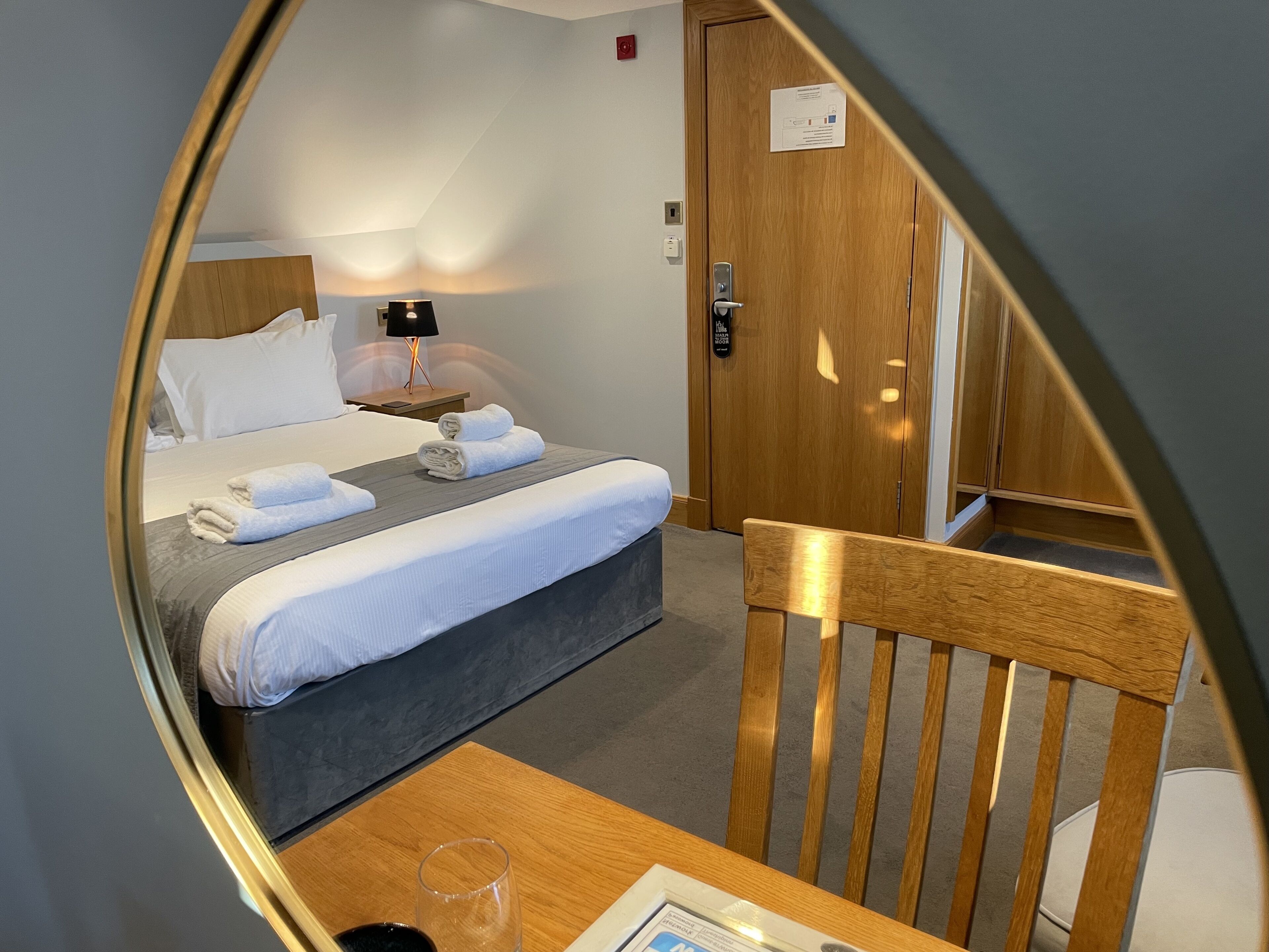 A hotel room with a bed, a nightstand with a lamp, a wooden chair, and a table with a glass and a menu.