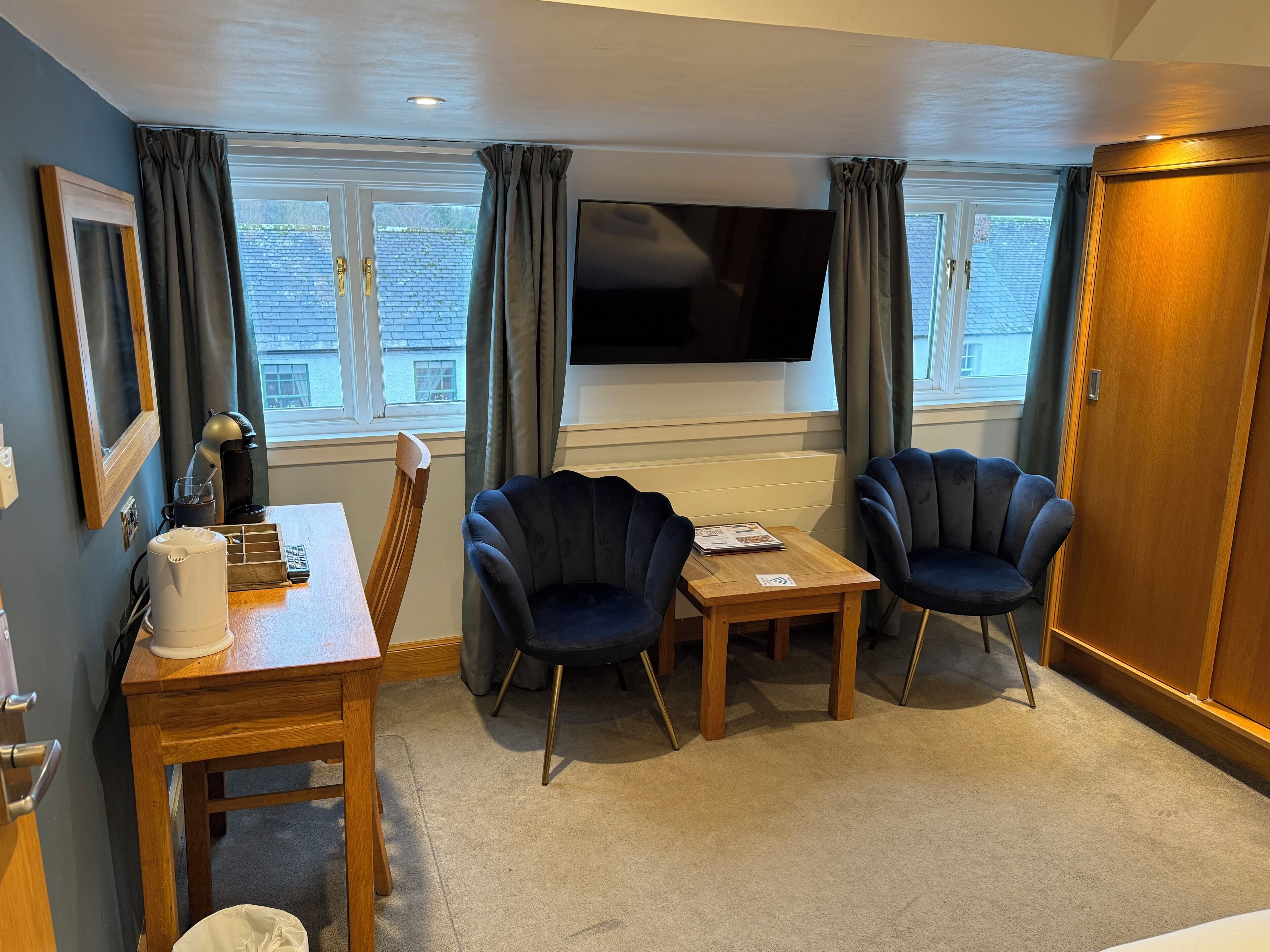 Ein Zimmer mit einem Holztisch, zwei dunklen Sesseln, einer Kaffeemaschine und einem Fernseher an der Wand.