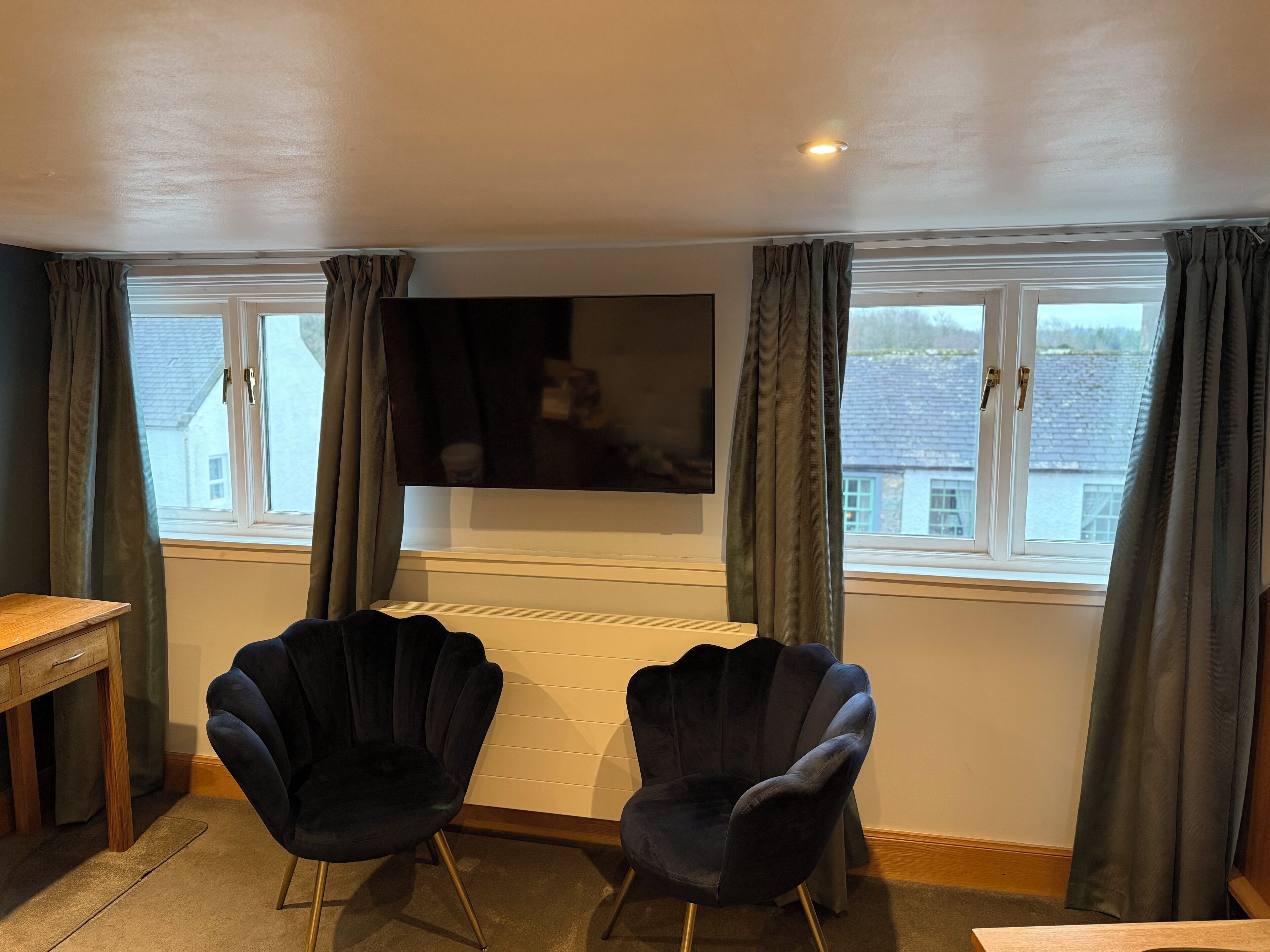 A living room with a flat-screen TV mounted on the wall, two dark armchairs, a wooden side table, and a view of a house through the window.