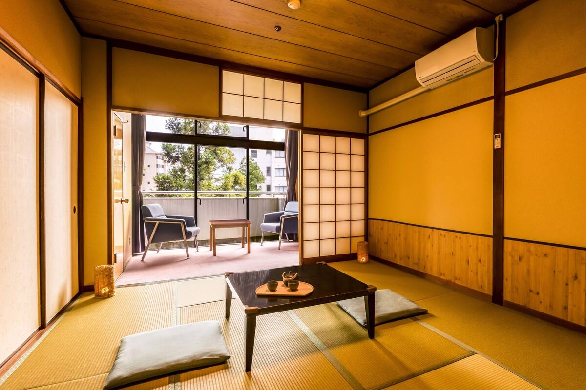 Ein traditioneller japanischer Raum mit Tatami-Böden, einem niedrigen Tisch mit Teeservice und Schiebetüren zu einem Sitzbereich im Freien.