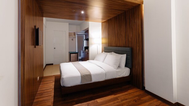 A bedroom with a wooden ceiling, a bed with white bedding, and a visible kitchen area in the background. - H Hotels - Metro North Uno (Quezon City)