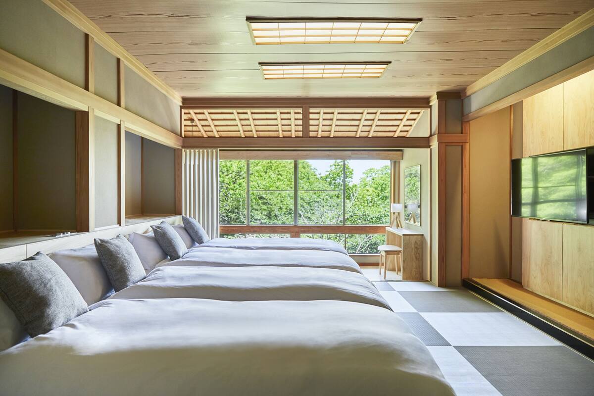A hotel room with a large bed, a TV mounted on the wall, and a view of the outdoors through large windows.