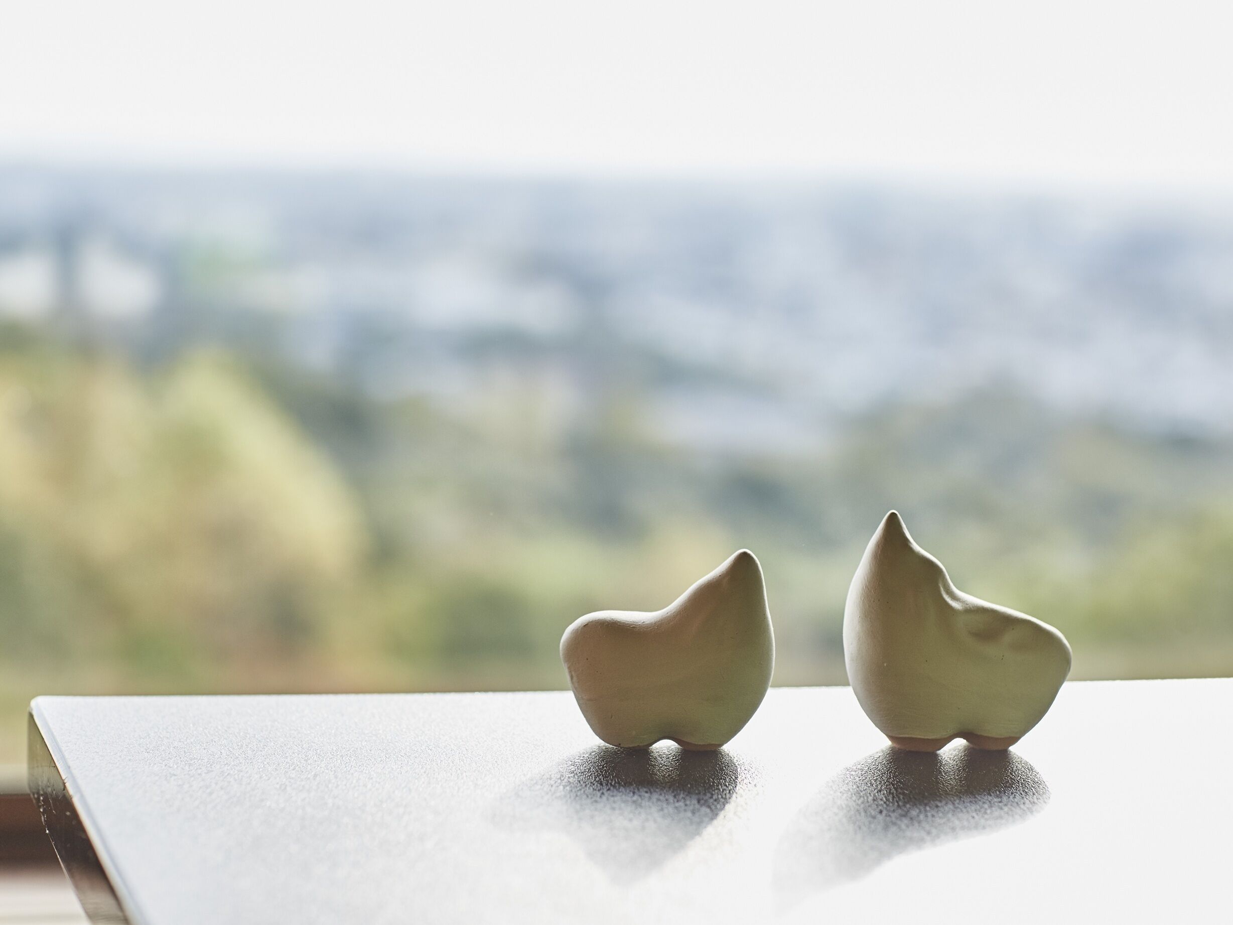 Two ceramic sculptures on a table with a blurred cityscape view in the background.