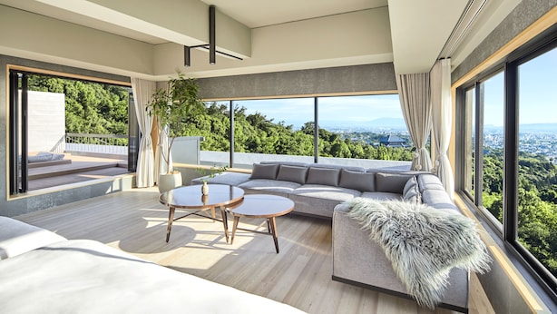 Una sala de estar moderna con un amplio sofá modular, dos mesas redondas de madera y vistas a un paisaje verde exuberante a través de grandes ventanales.