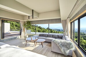 Un salon moderne doté d’un grand canapé d’angle, de deux petites tables rondes en bois et offrant une vue sur un paysage verdoyant grâce à de grandes fenêtres.