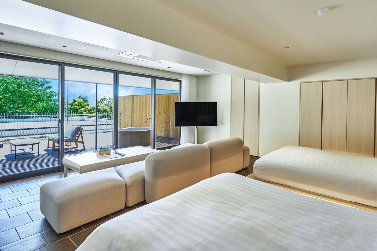 A modern hotel room with a large bed, a sofa, a TV, and a balcony with a view of greenery.