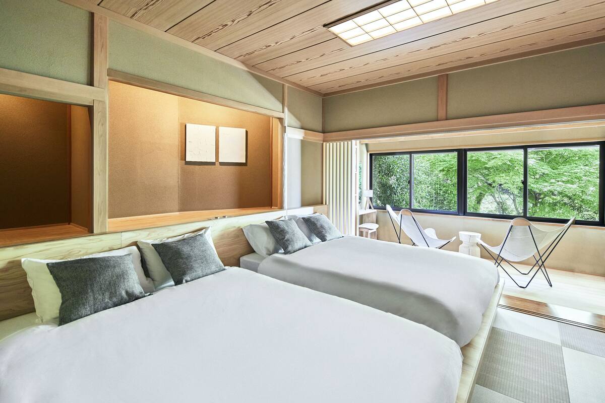 A hotel room with two beds, a large window with a view of greenery, a wooden ceiling, and a small seating area with a chair and table.