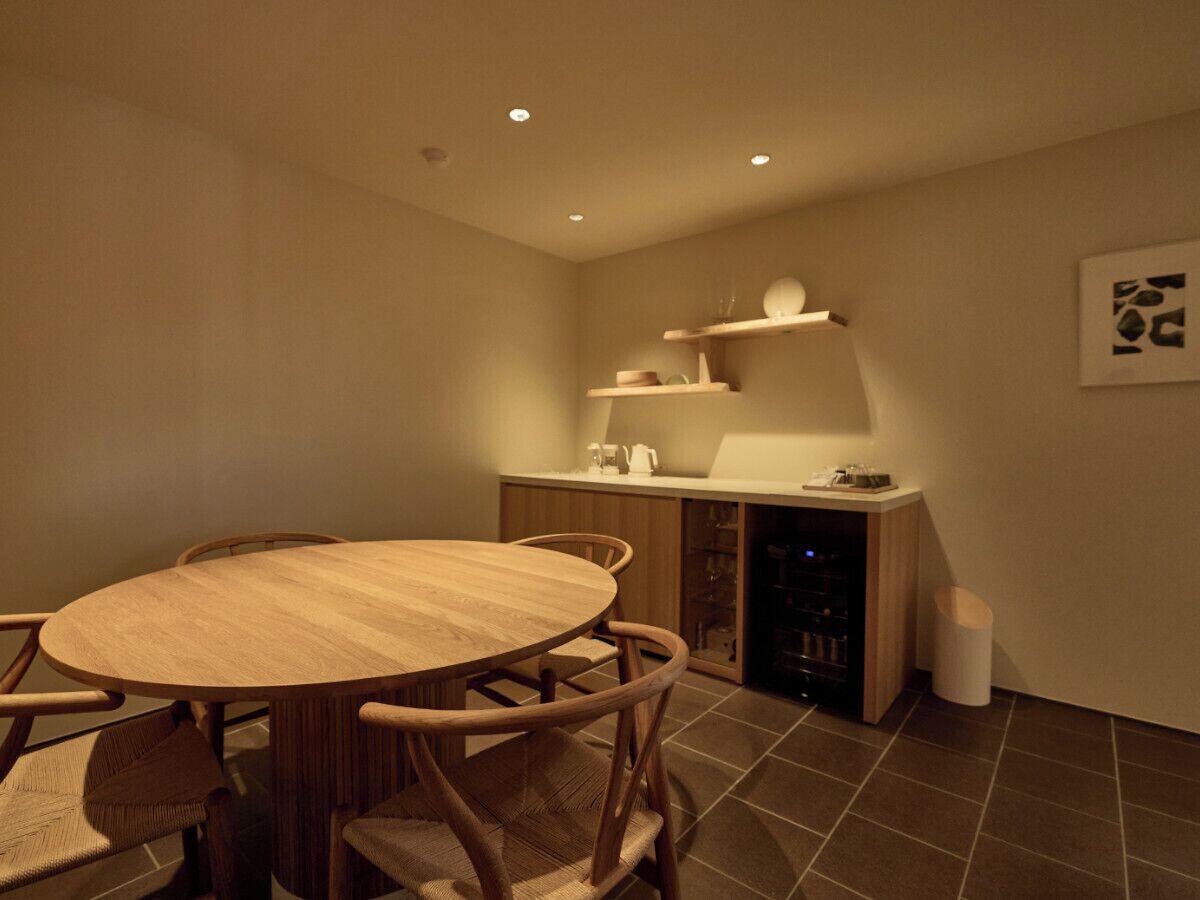 Un coin repas compact avec une table ronde en bois et des chaises, un meuble à vin intégré et un petit espace cuisine doté d’un plan de travail et d’étagères.