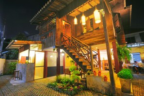 A traditional wooden building with a staircase leading to a second floor, a small outdoor seating area with stools, and potted plants. - Manon Hostel (Chiang Mai)