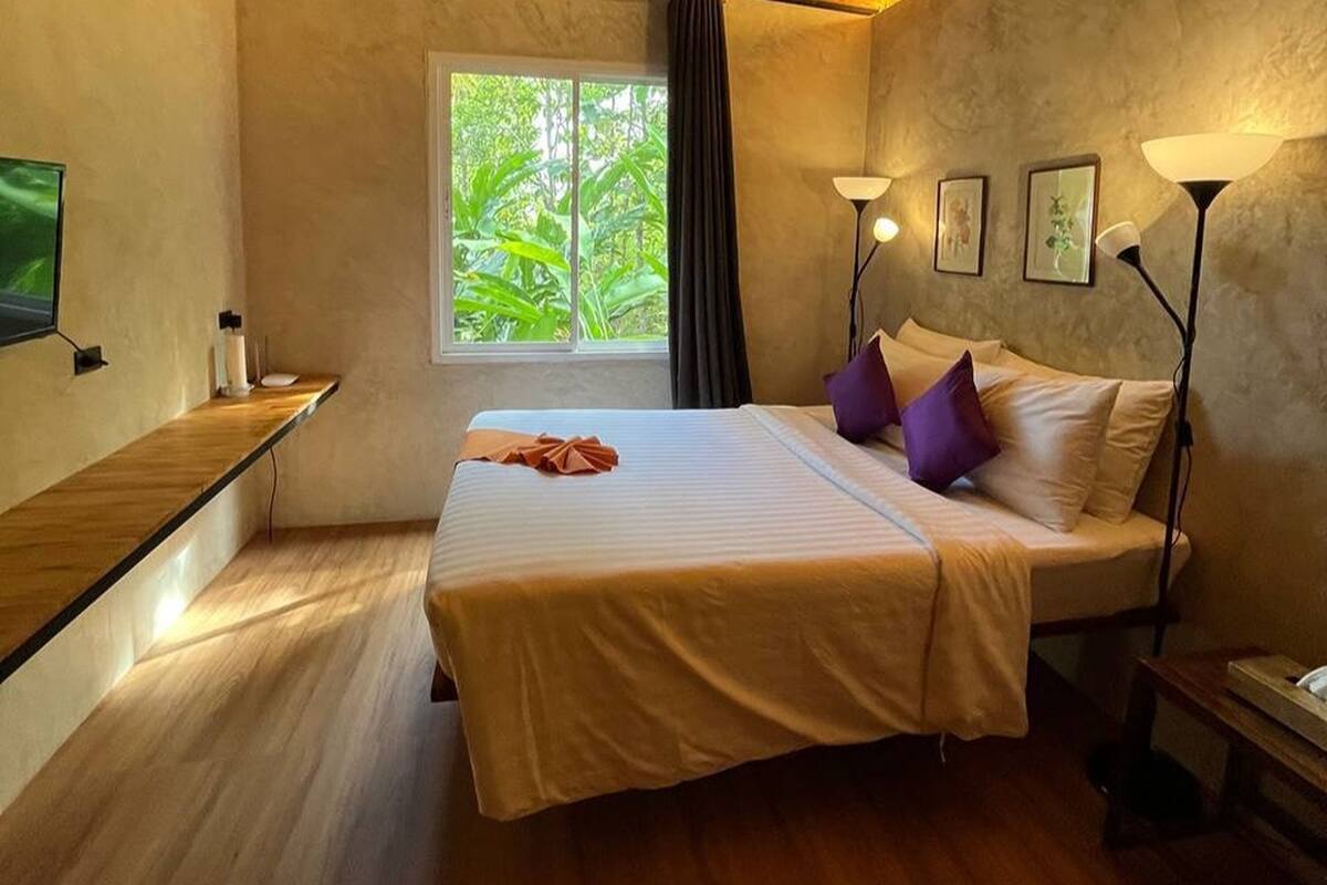 A bedroom with a bed, a window with a view of greenery, a bedside table with a lamp, and a TV mounted on the wall.