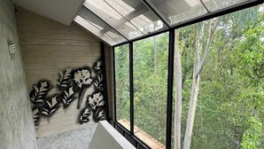 Une pièce dotée d’une grande fenêtre donnant sur une forêt, d’un mur en béton orné d’un motif de feuilles et d’un plafond en pente avec des lucarnes.