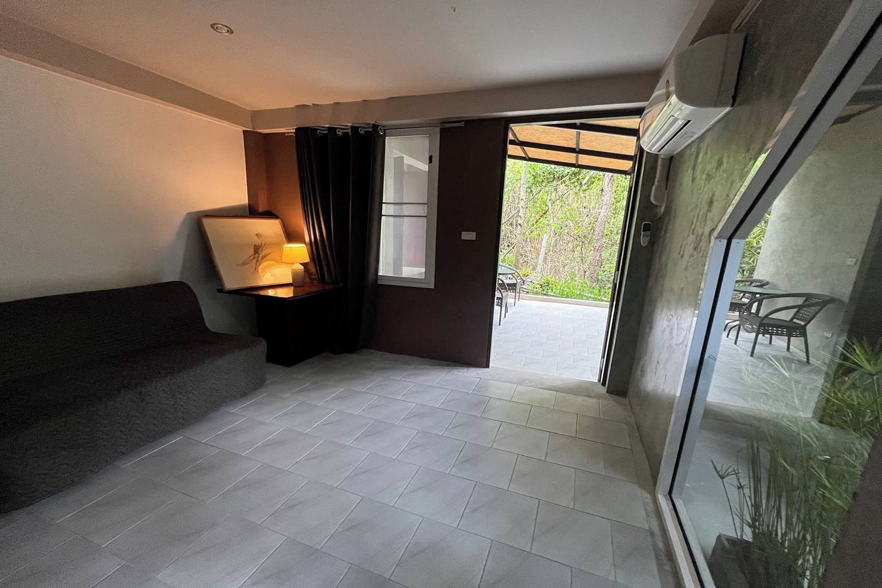 A modern room with a grey sofa, a small table with a lamp, a sliding glass door leading to an outdoor area with a table and chairs.