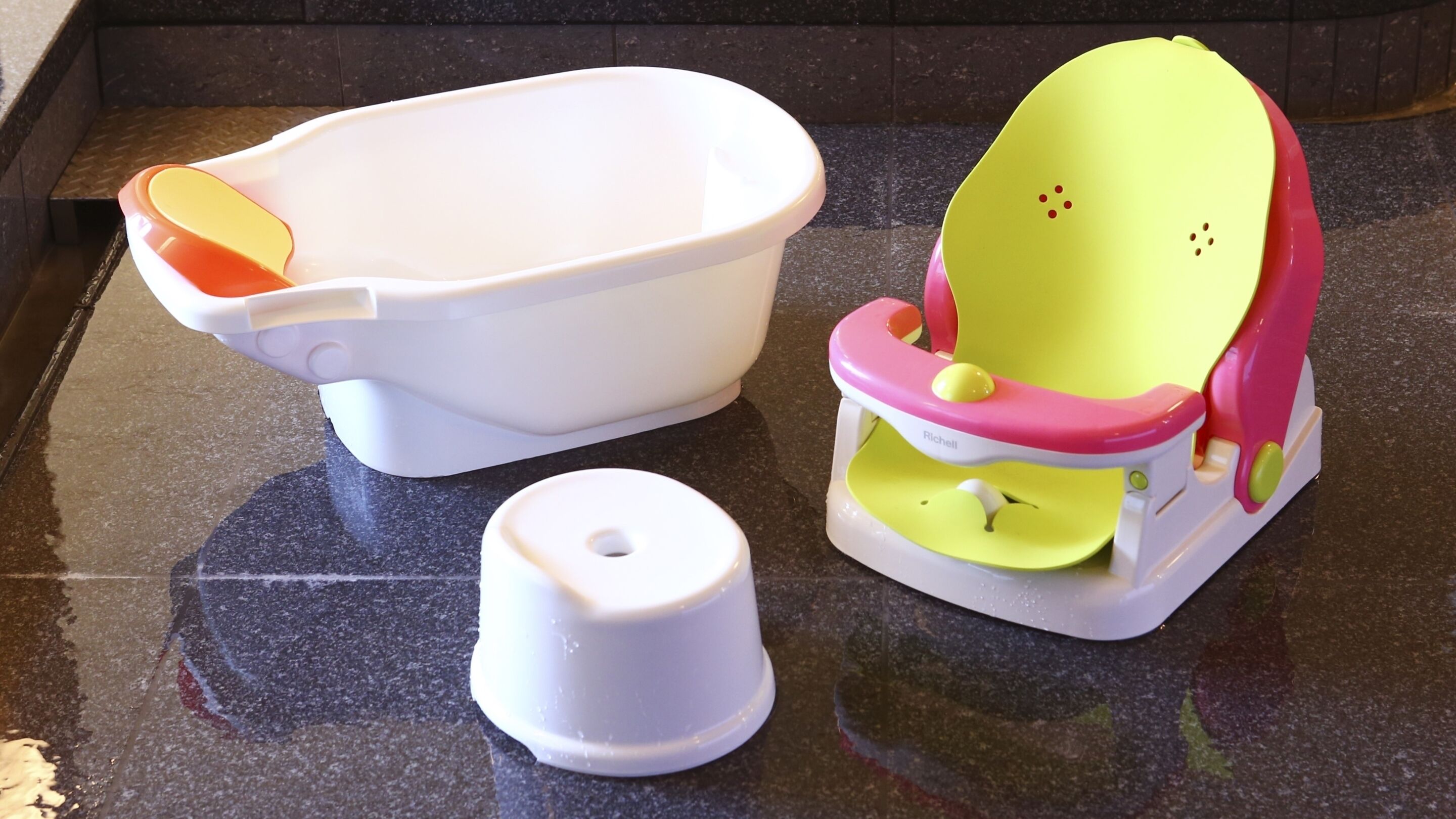 Una encimera de cocina con un lavabo de plástico blanco, una trona rosa y amarilla, y un recipiente de plástico blanco.