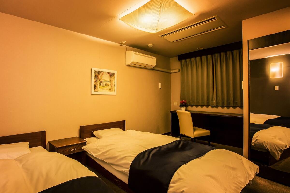 A hotel room with two beds, a desk, a chair, an air conditioning unit, a painting, and a mirror.