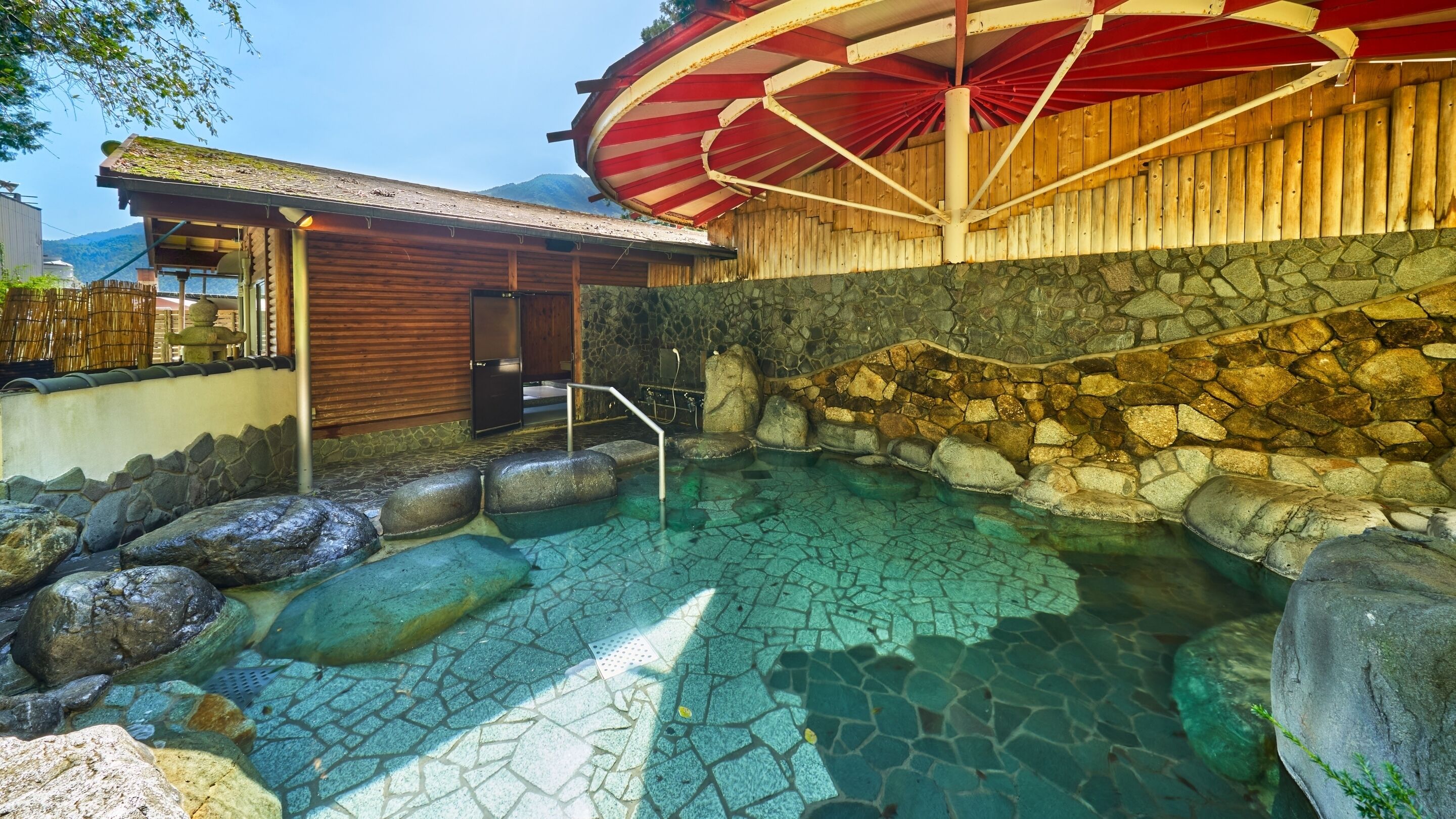 Una piscina de piedra con rocas, una estructura de madera y una sombrilla roja.