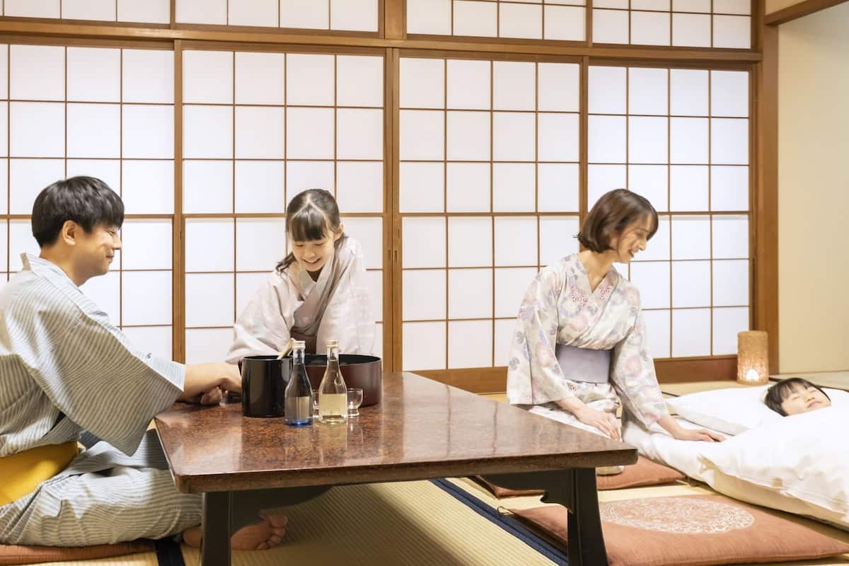 A tatami mat room with a low table, three people sitting, and a person lying down.