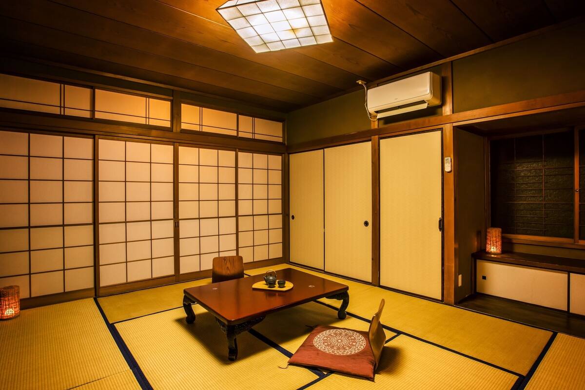 A traditional Japanese room with tatami flooring, a low table, a cushion, sliding doors, and a window with a view.