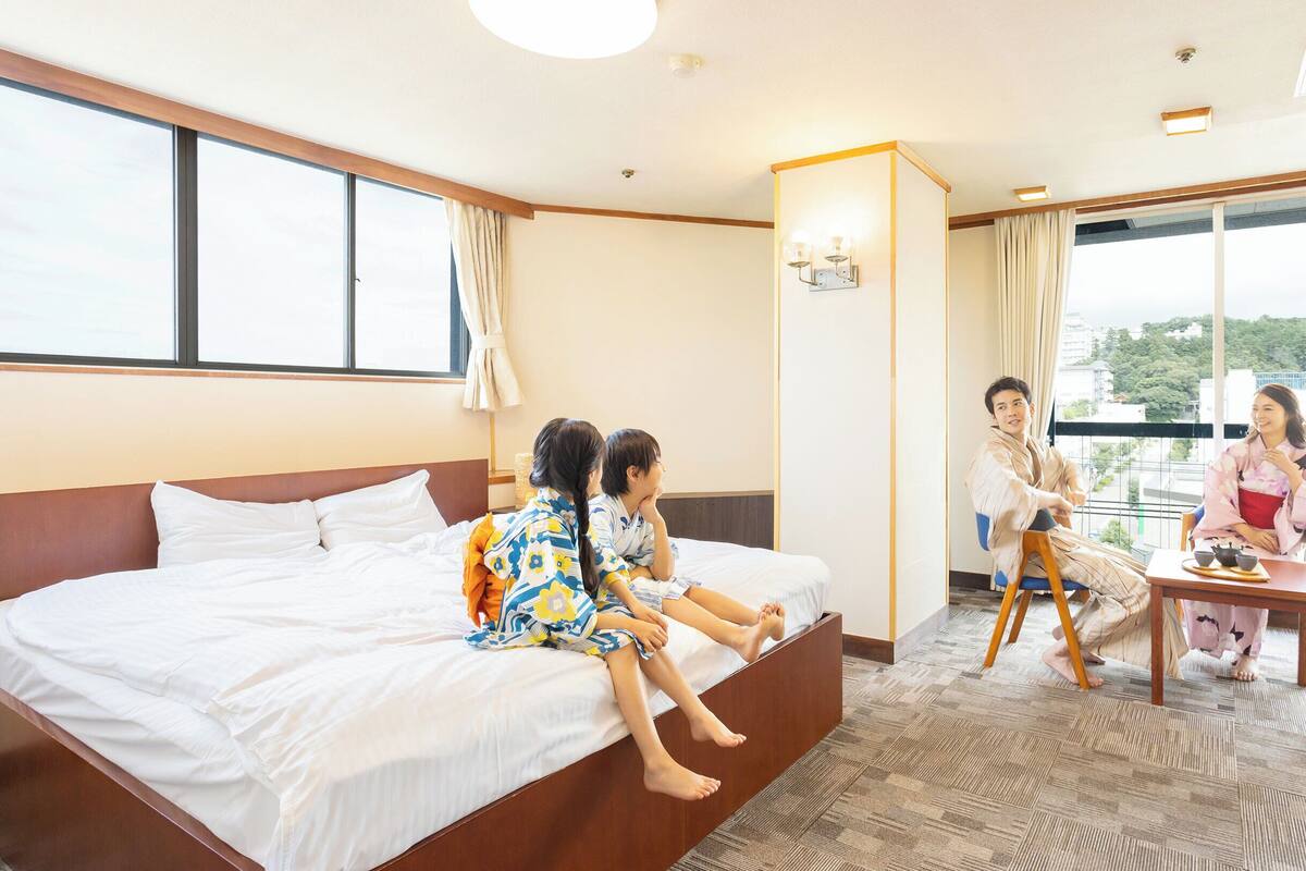 A hotel room with a large bed, a sitting area with a table, and a view of buildings through large windows.