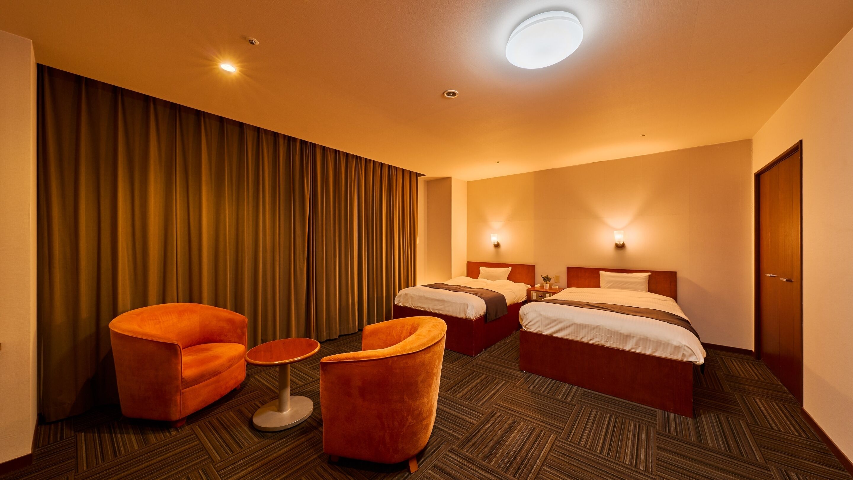 Ein Hotelzimmer mit zwei Betten, einem kleinen Tisch, zwei orangenen Sesseln und einem großen Fenster mit Vorhängen.