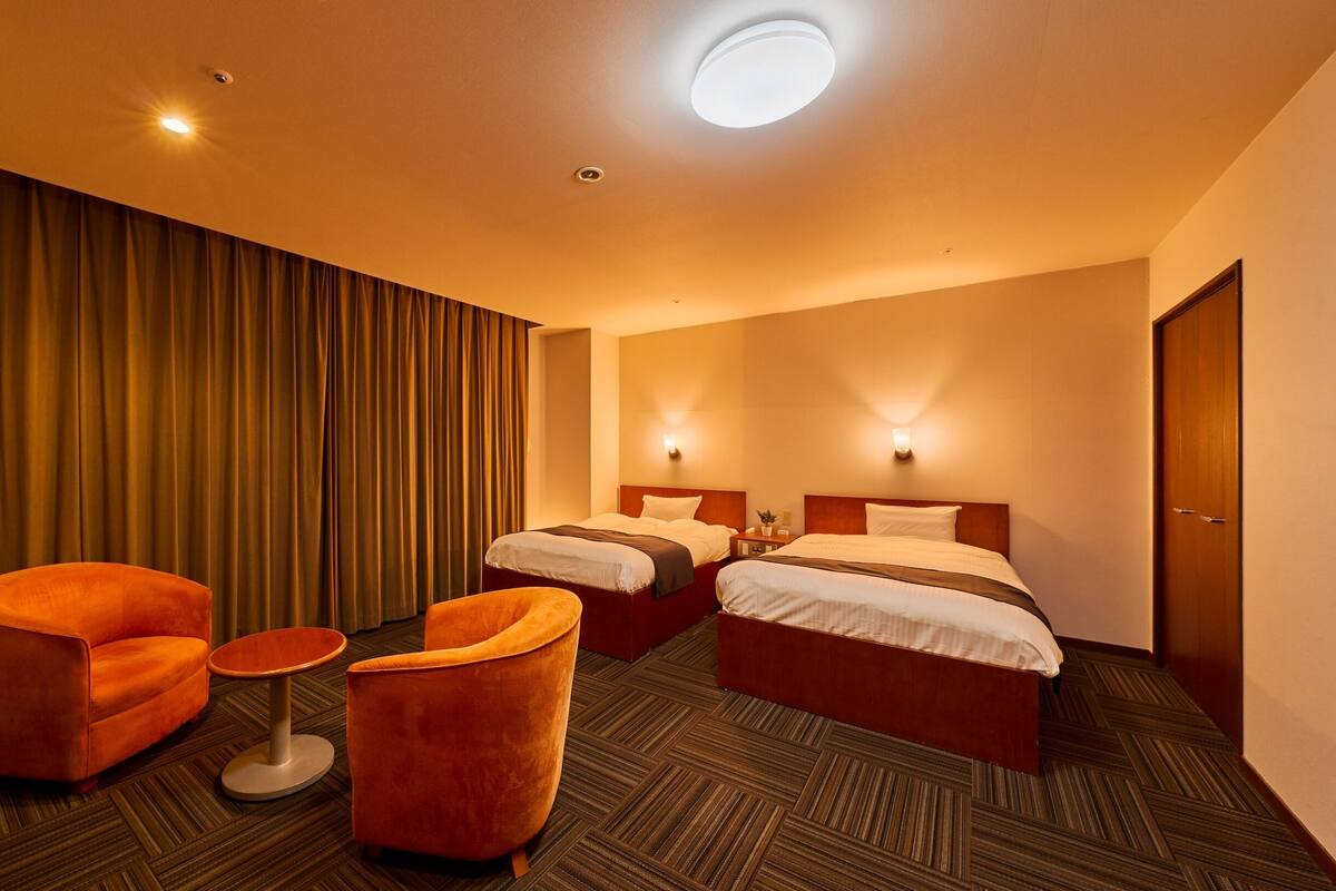A hotel room with two beds, a small table, two orange armchairs, and a large window with curtains.