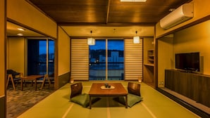 Une pièce de style japonais traditionnel, avec un sol recouvert de tatamis, une table basse avec des coussins, un téléviseur et une vue sur le paysage urbain à travers de grandes fenêtres.