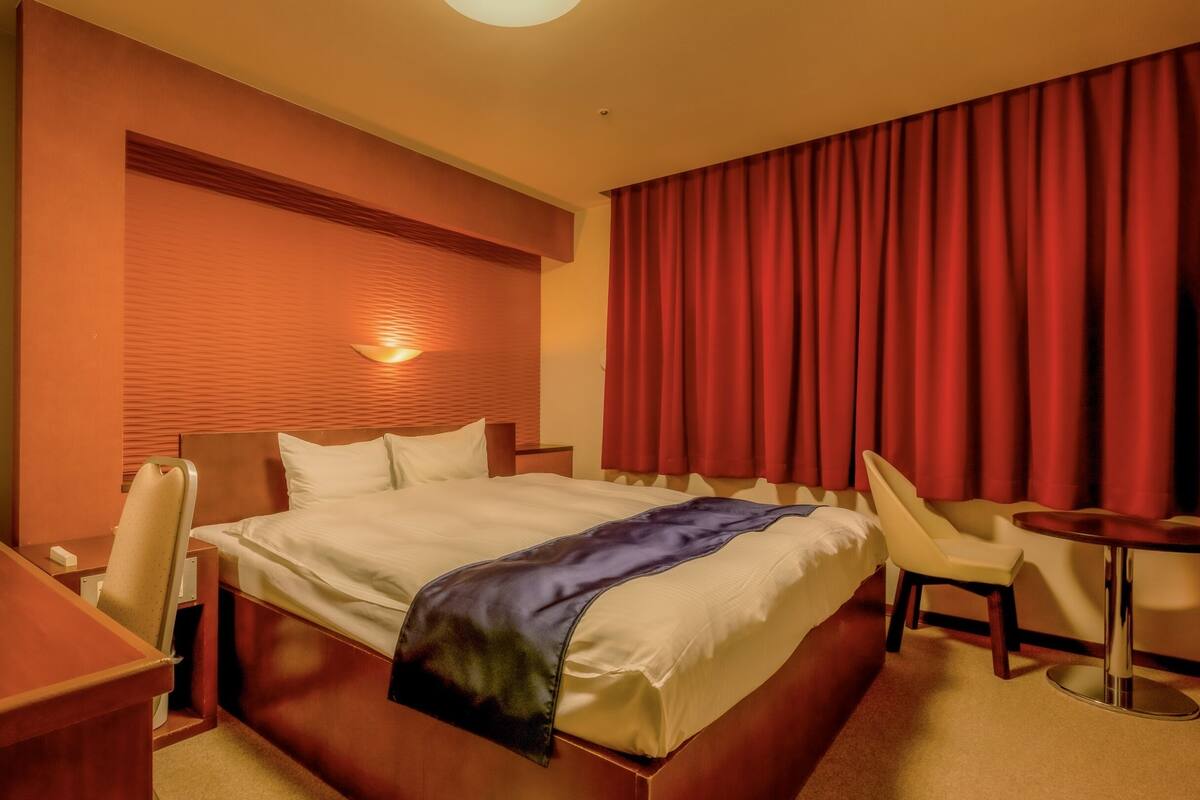 A hotel room with a bed, a desk, a chair, a small table, and a red curtain.