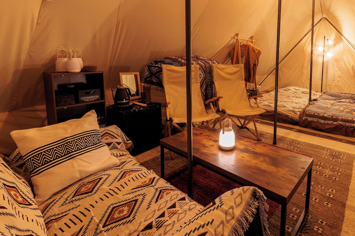 A tented accommodation with a bed, a table with a light, a chair, and a small shelf with items.