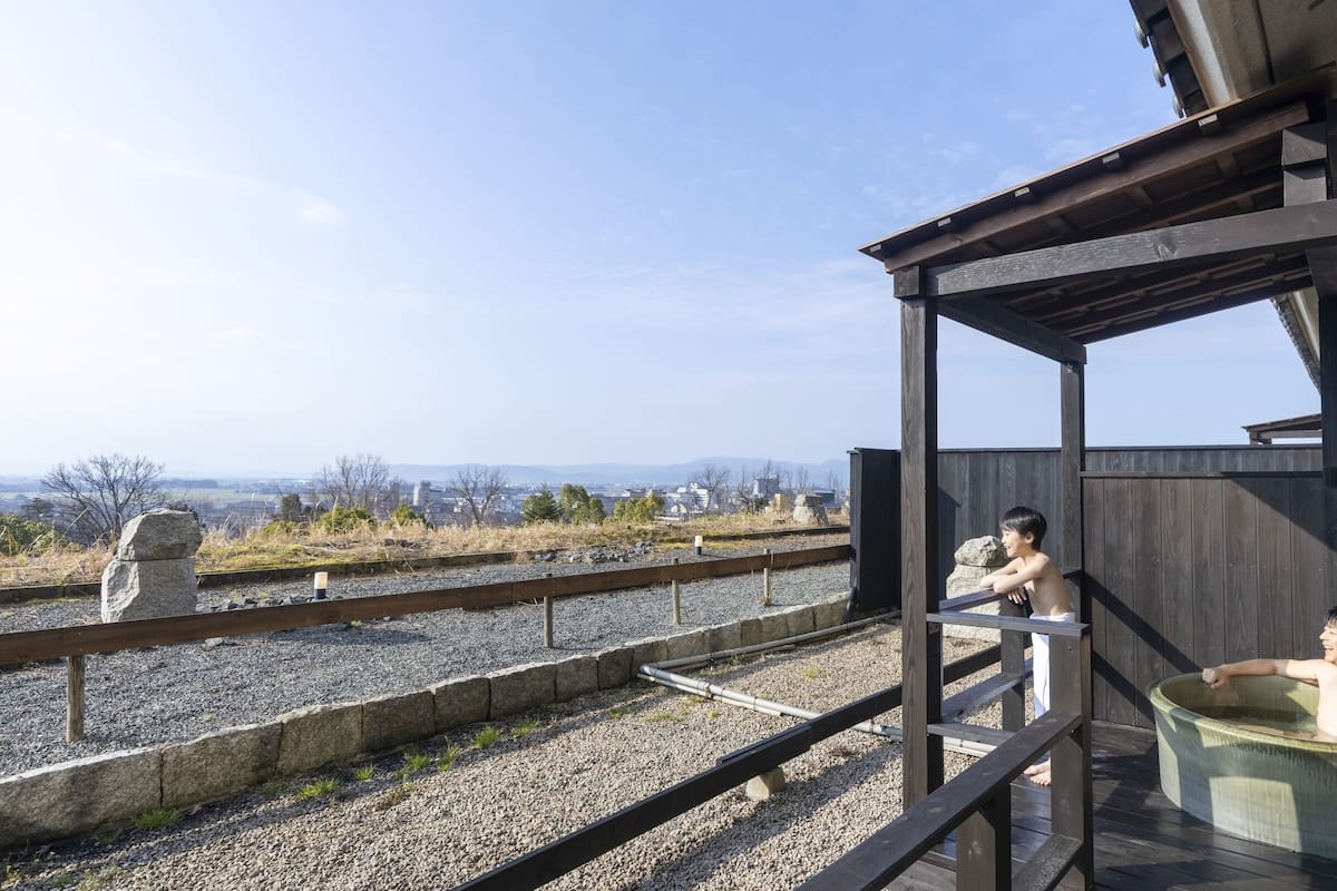 A wooden deck with a stone wall, a wooden fence, and a view of distant buildings and mountains.